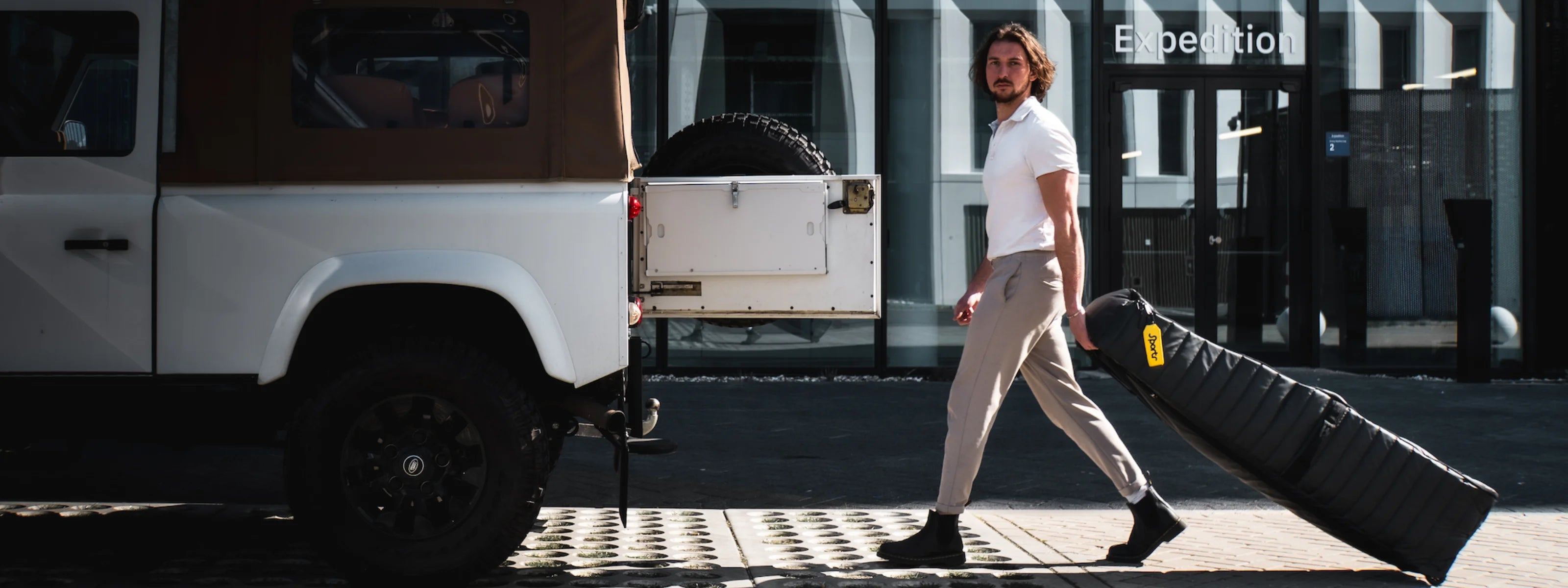 Person walking towards a white Landrover with an open back door, carrying a Sportr Golf Travel Bag. 